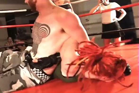 Black-haired dominatrix wields wooden paddle over kneeling opponent in red-walled ring on breastful.com